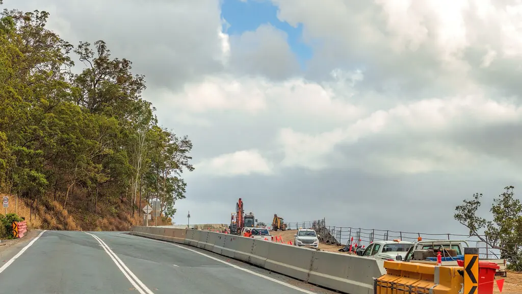 A winding road passes through a construction zone with traffic barriers, machinery, and parked vehicles. Trees line the left side, and the sky is partly cloudy.