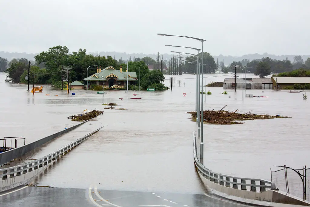 A flooded street with water covering the road and surrounding buildings; debris floats in the water and only the roofs and tops of some structures and trees are visible under a gray, overcast sky.