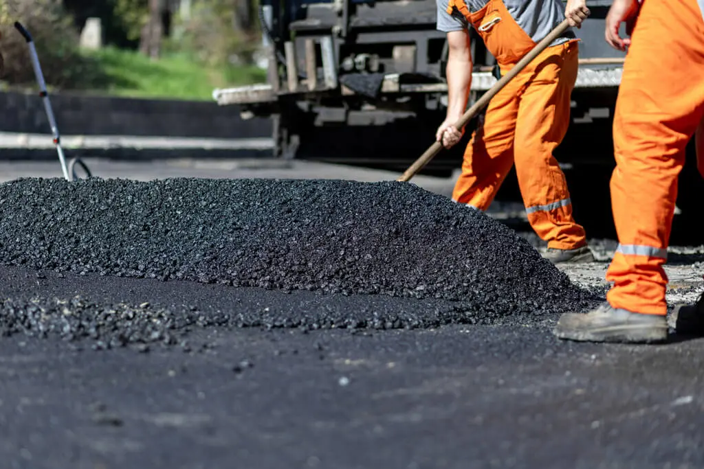 Two workers in orange uniforms spread fresh asphalt on a road with shovels. A mound of black asphalt is in the foreground, and roadwork equipment is visible in the background.