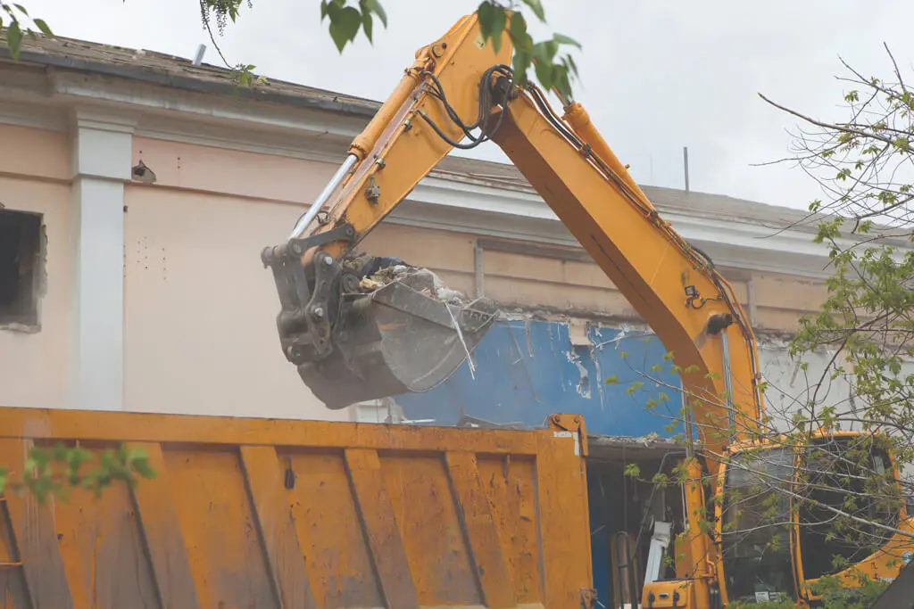 A yellow excavator loads debris into a large orange dump truck in front of a partially demolished building, with dust and damaged walls visible. Green branches frame the scene.
