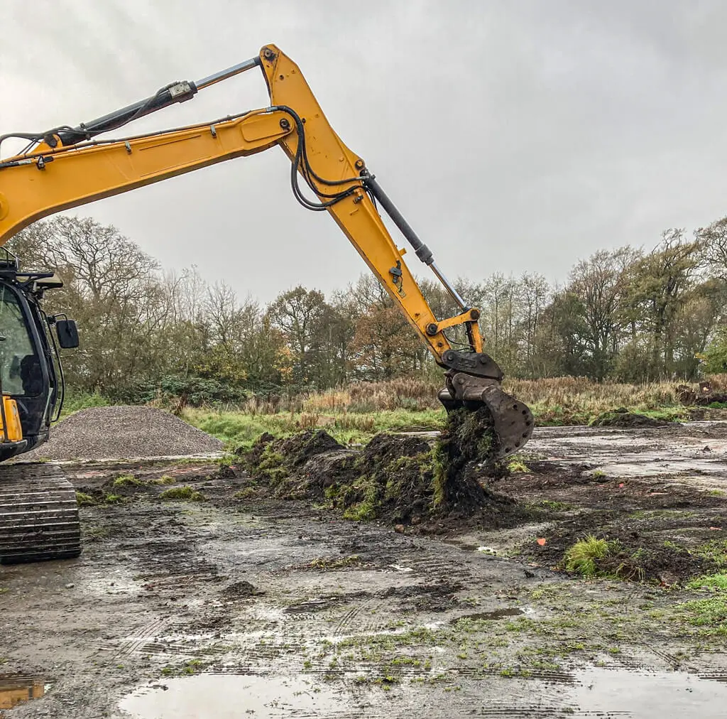 A yellow excavator is digging up soil and grass on a muddy construction site with trees and cloudy sky in the background.