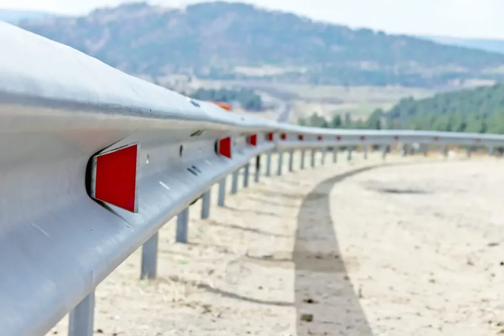 A close-up view of a metal highway guardrail with red reflectors, curving along a road with a blurred mountain and trees in the background.