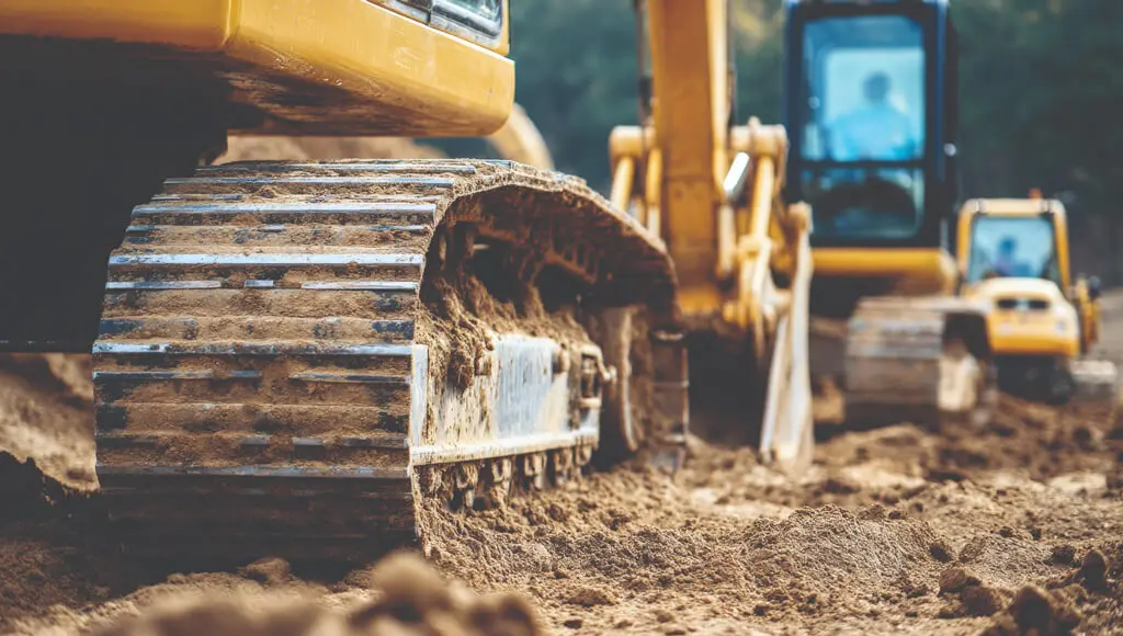 Close-up view of a construction vehicle’s metal tracks on sandy soil, with another excavator in the background. The scene highlights bout MIH’s operator-led contracting and commitment to a strong safety culture on-site.