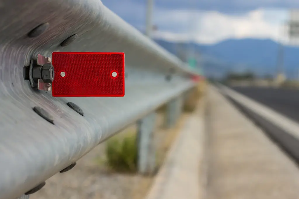 Close-up of a metal roadside guardrail with a small red reflective marker attached, emphasizing safety culture along the paved road. The blurred background reveals mountains and an overcast sky.